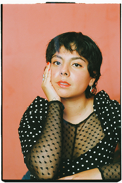 Peach background, a mexican woman with short black hair, dressed in black with white spots.