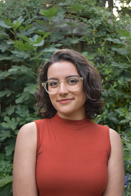 Aysha Zackria, a person with short curly hair, glasses, and a rust-colored sleeveless top, standing in front of green foliage.