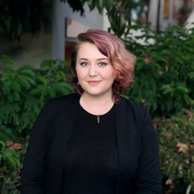 A non-binary person with pink hair wearing all black. Green foliage in the background.