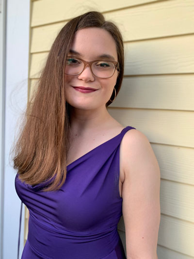 A white woman with long brown hair and glasses, wearing a purple dress. Photo credit: Lauren Backus 