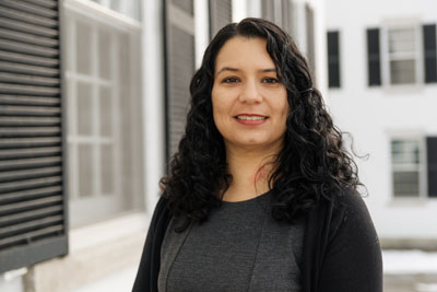 A woman with medium skin tone and long, curly black hair is standing outdoors in front of a white building with black shutters. She is wearing a dark gray dress with a black cardigan over it.