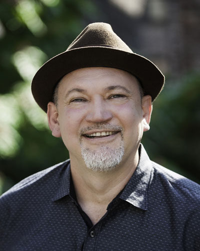   A white Latino man with a short gray goatee wearing a brown fedora hat and a gray buttoned down shirt. Photo credit: Michael Palma. 