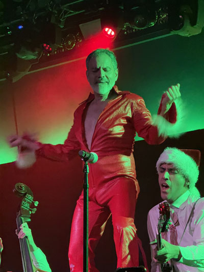 El Vez, a handsome older Chicano man, dances atop a stand-up bass wearing a fur-trimmed, red vinyl jumpsuit. 