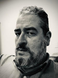 The image is a close-up, black-and-white portrait of a middle-aged man with a strong, serious expression. He has a broad face with prominent features, including a full beard and mustache that are peppered with gray. His hair is short, wavy, and also shows signs of graying, particularly at the temples and top. 