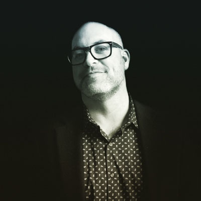 A black and white photo of a 50-year old man with glasses and a shaved head.