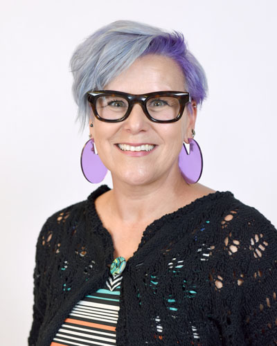 A woman with purple hair and glasses wearing a colorful striped dress. 