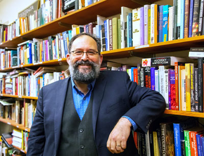 Michael is a black-haired, black-bearded man with glasses smiling while leaning against a bookcase.
