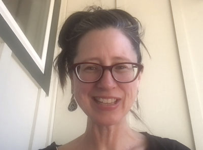 A smiling White woman with glasses and a ponytail, wearing dangling earrings
