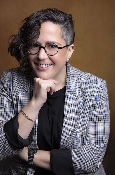  A woman with round black glasses, short hair with one side shaved and the other curly, a smile, and an outfit consisting of a checkered blazer and a black shirt. Photo credit: Odette Villareal