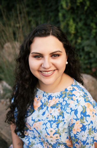 A mixed-race Ojibwe/White woman with long curly brown hair wearing a blue floral dress. Photo Credit: Stephanie Tumbleson