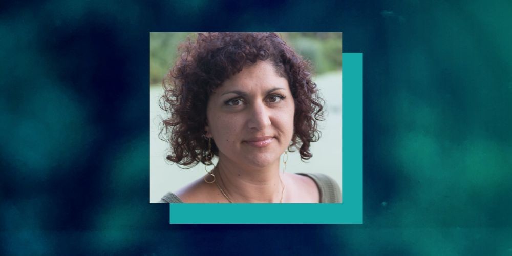 A photo of Meropi Peponides with curly hair, wearing a green top and a necklace, against a blurred outdoor background.