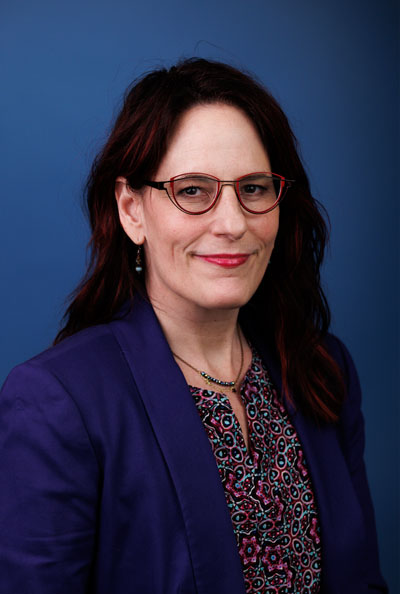 Una mujer blanca con cabello castaño oscuro y gafas rojas con una chaqueta violeta.