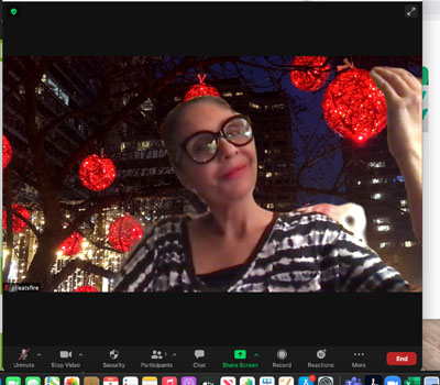 Una mujer de piel morena clara y cabello castaño sostiene una gran luz navideña roja y brillante. Lleva el cabello recogido en un moño. Lleva lentes de montura grande y lápiz labial rojo. Sonríe con curiosidad.