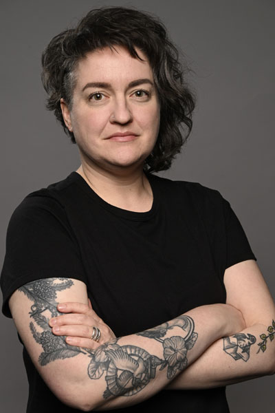 A white person with asymmetrical, dark curly hair and tattoos on their arms.