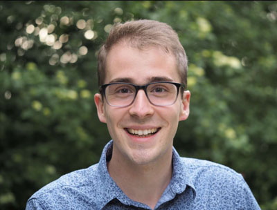 Un hombre blanco con cabello castaño que viste una camisa azul abotonada y lentes.