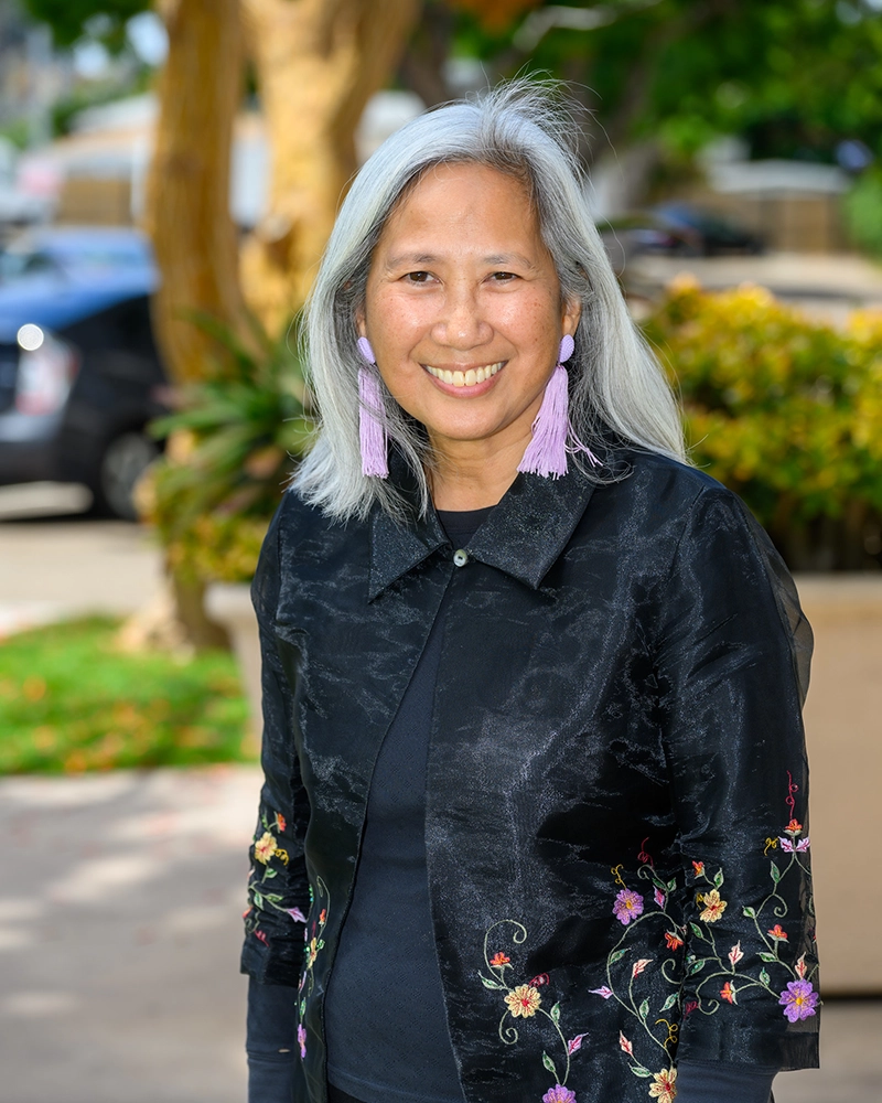 Thelma Virata de Castro smiling, wearing purple tassel earrings and a black jacket with floral embroidery.
