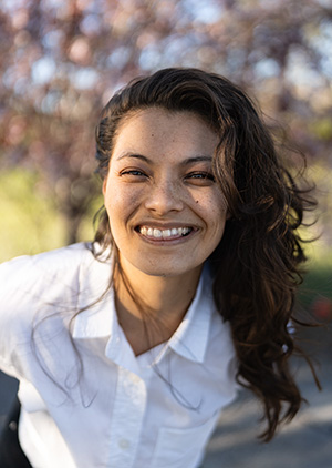 Una persona filipina de raza mixta, con cabello castaño ondulado y pecas, viste una camisa blanca con cuello y sonríe contra un fondo borroso de flores de cerezo.