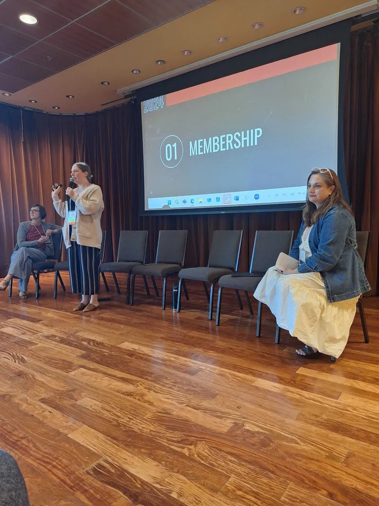 A person speaks into a microphone while standing on a stage beside a large projection screen. Two others sit nearby, one in a chair to the left and one on the right, while several empty chairs line the stage.