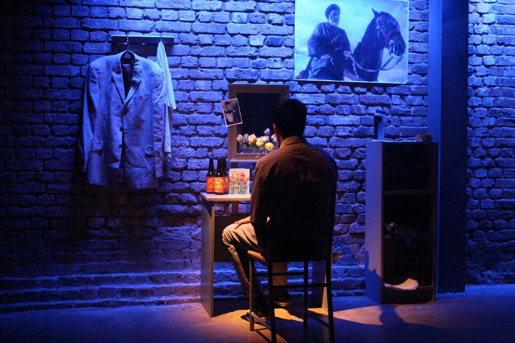 A performer seated at a desk facing a brick wall in Love, Bombs and Apples