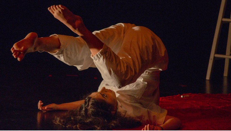 A performer lying on a red rug onstage in Regarding Antigone